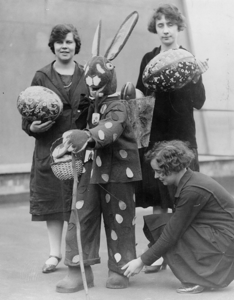 An Easter bunny carries easter eggs in a basket on his back while one woman adjusts his turn-ups and two others carry large easter eggs, circa 1930.
